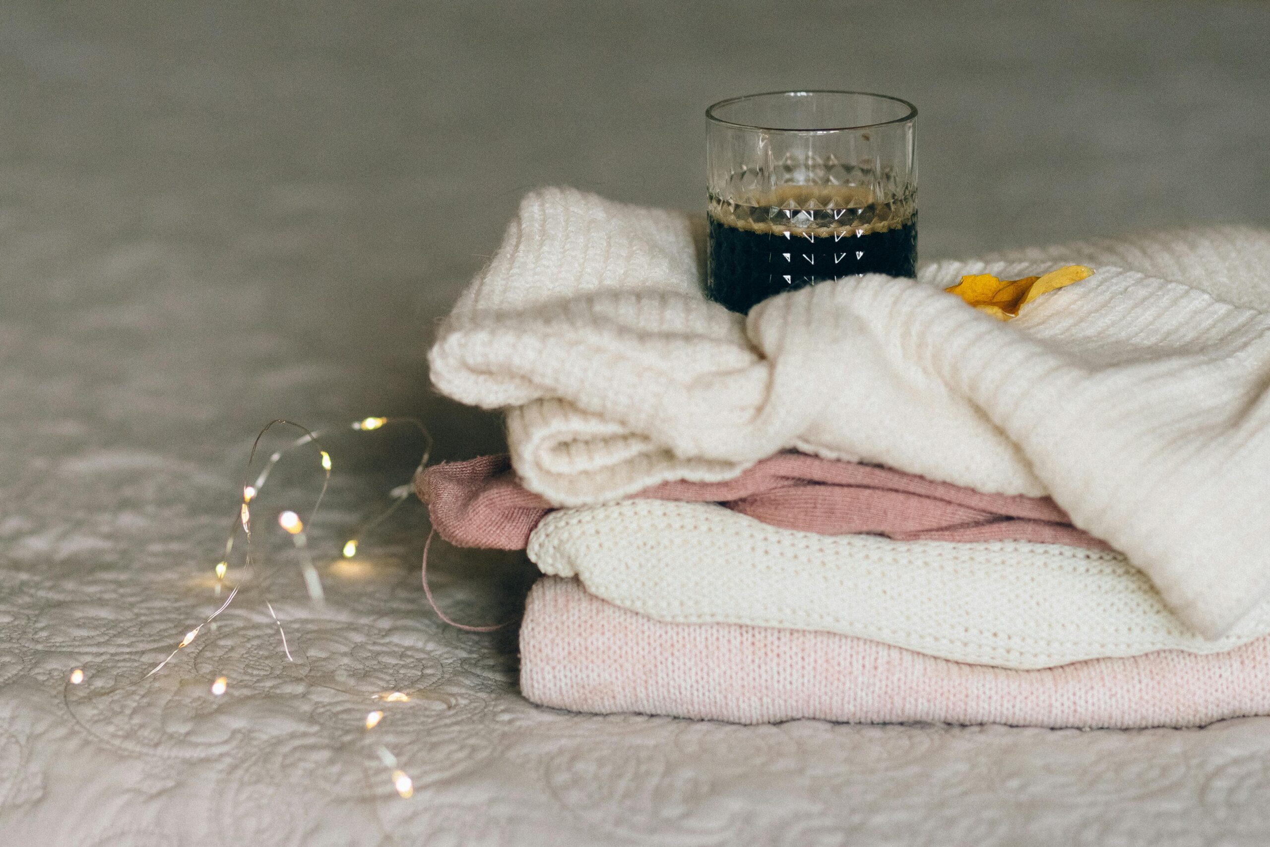 Stack of knitted sweaters with a warm drink beside cozy fairy lights.
